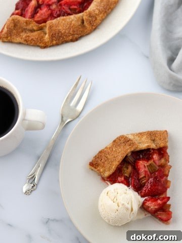 A slice of strawberry rhubarb galette with a scoop of ice cream and a fork and a cup of coffee