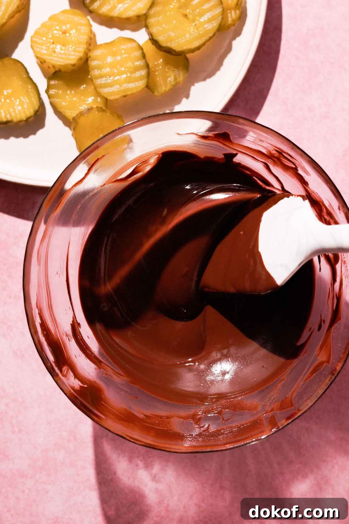 melted chocolate in glass bowl with rubber spatula and sliced pickles on the side