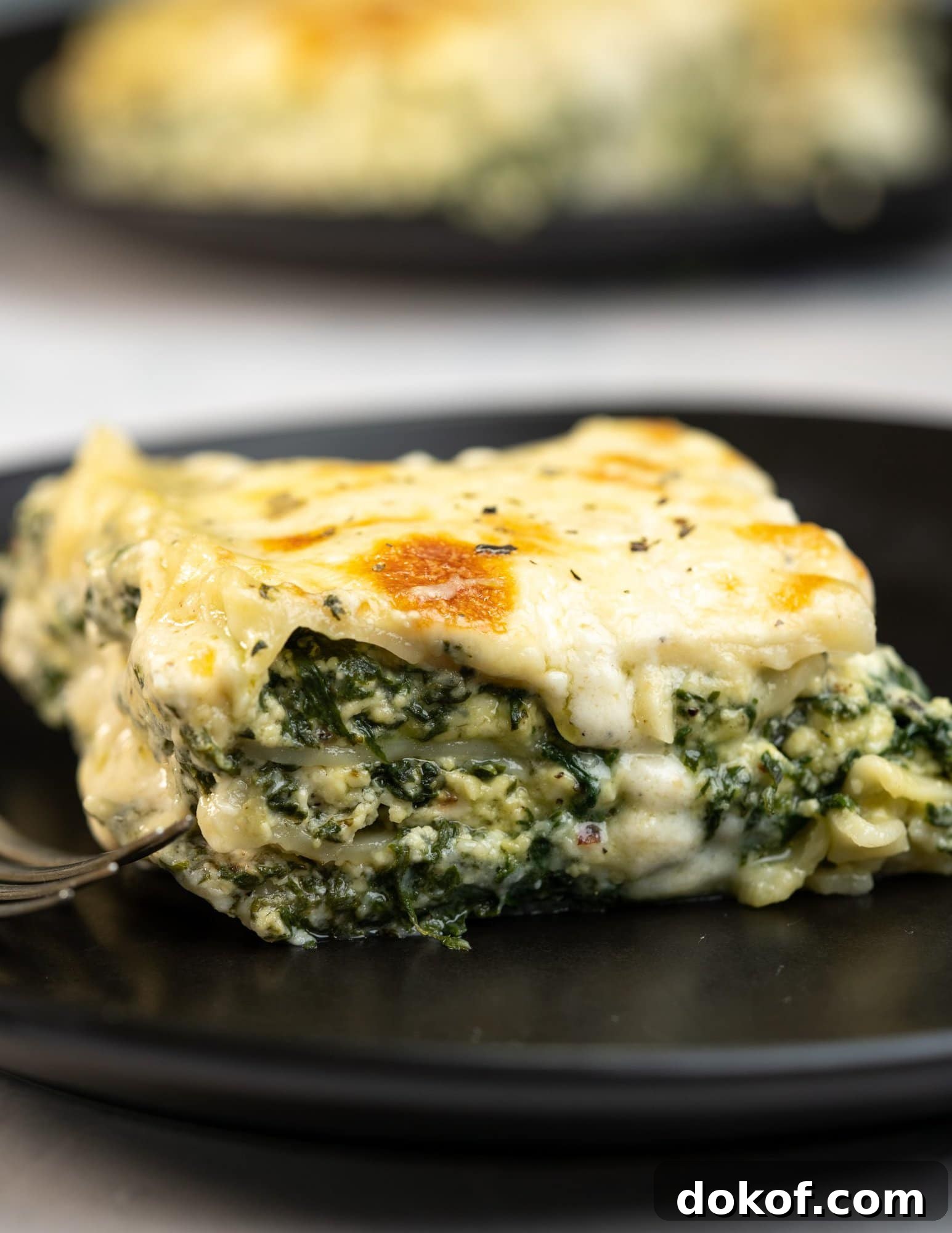 A slice of spinach lasagna with cheese oozing out of the layers and served on a black dining plate.