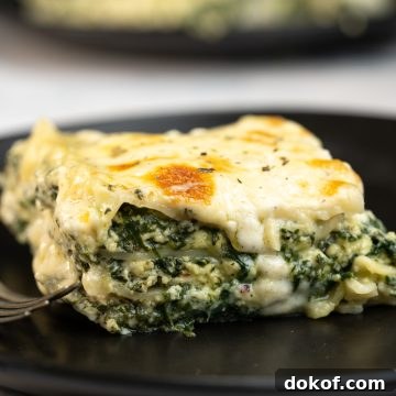 A slice of spinach lasagna with cheese oozing out of the layers and served on a black dining plate.