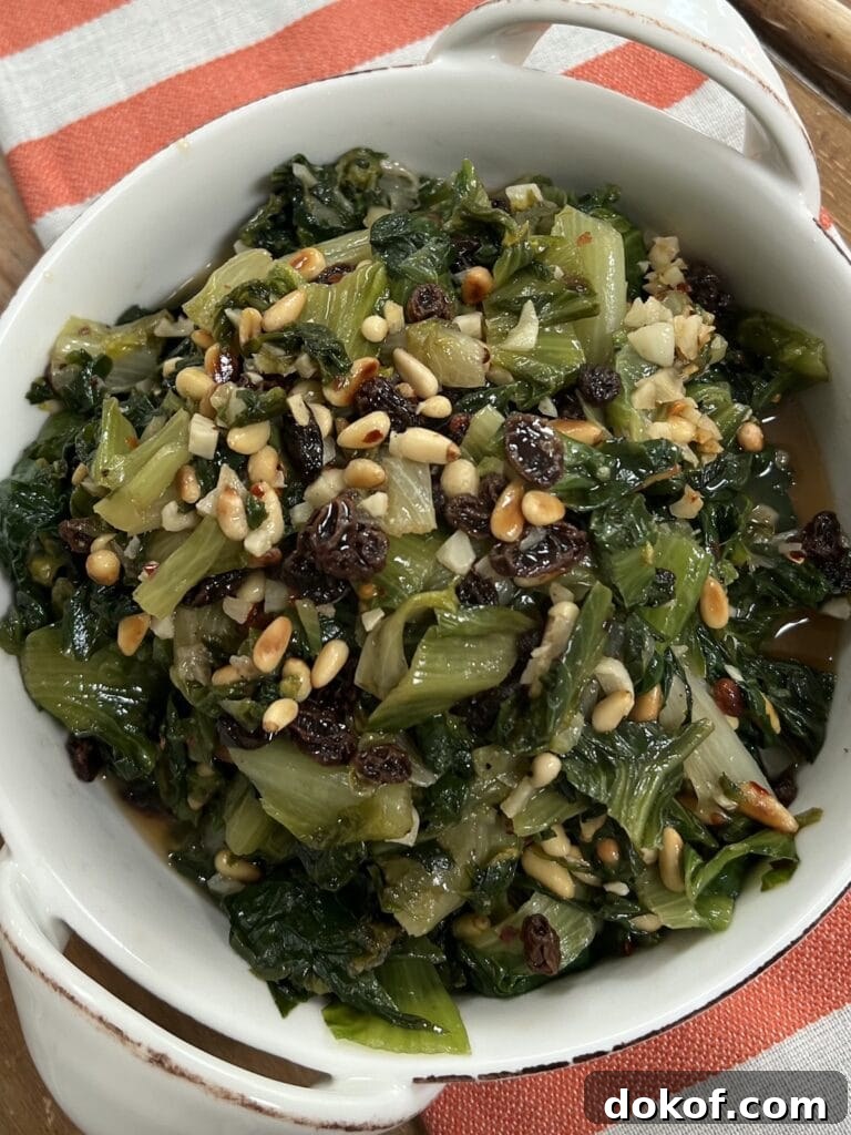Sautéed Italian Escarole with pine nuts and raisins in white dish over decorative towel.
