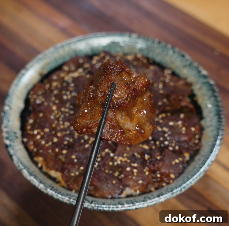Chopsticks are holding a saucy slice of bulgogi beef above a rice bowl, highlighting the juicy texture and rich glaze. The bowl below is packed with more beef slices and sesame seeds.