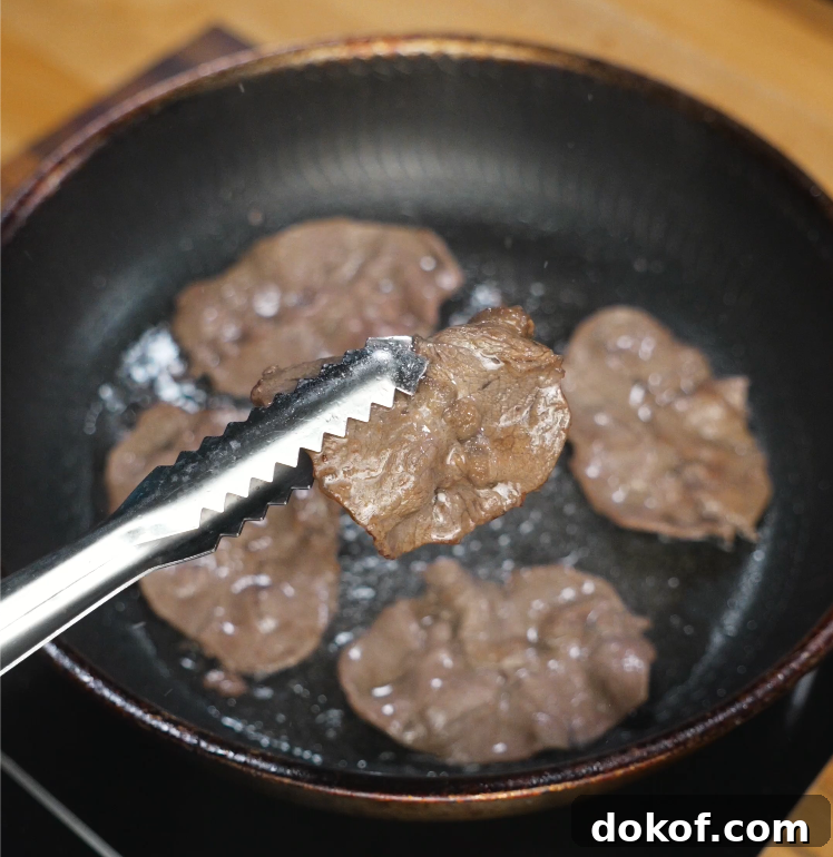 Slices of thinly cut beef are being seared in a hot pan, with tongs lifting one piece to show the browned surface. The meat is just beginning to caramelize for maximum flavor.