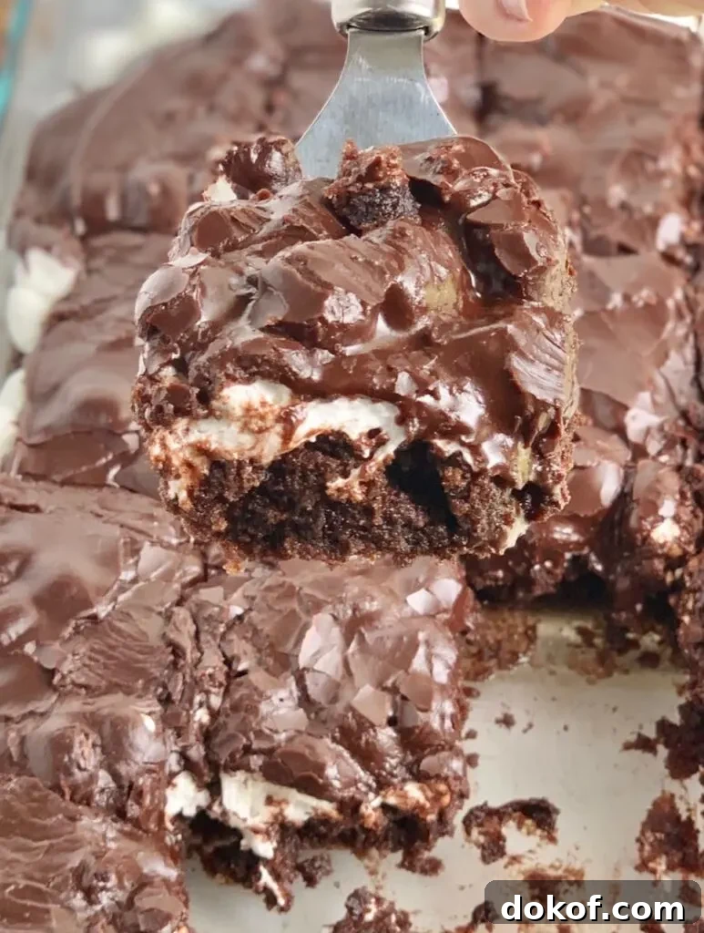 A glass baking dish filled with a dark brown chocolate cake followed by a white layer of mini marshmallows and topped with dark brown chocolate frosting. The bars have been cut into squares and some are missing from the pan. A spatula has lifted a slice out of the pan.