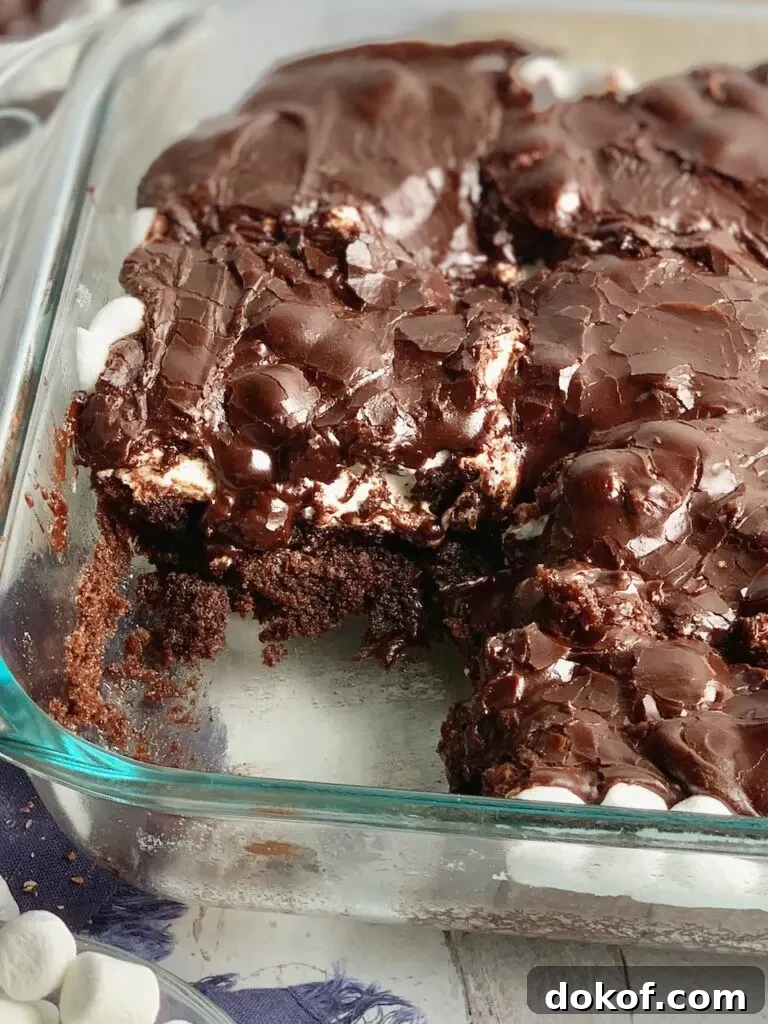 A glass baking dish filled with a dark brown chocolate cake followed by a white layer of mini marshmallows and topped with dark brown chocolate frosting. The bars have been cut into squares and some are missing from the pan.