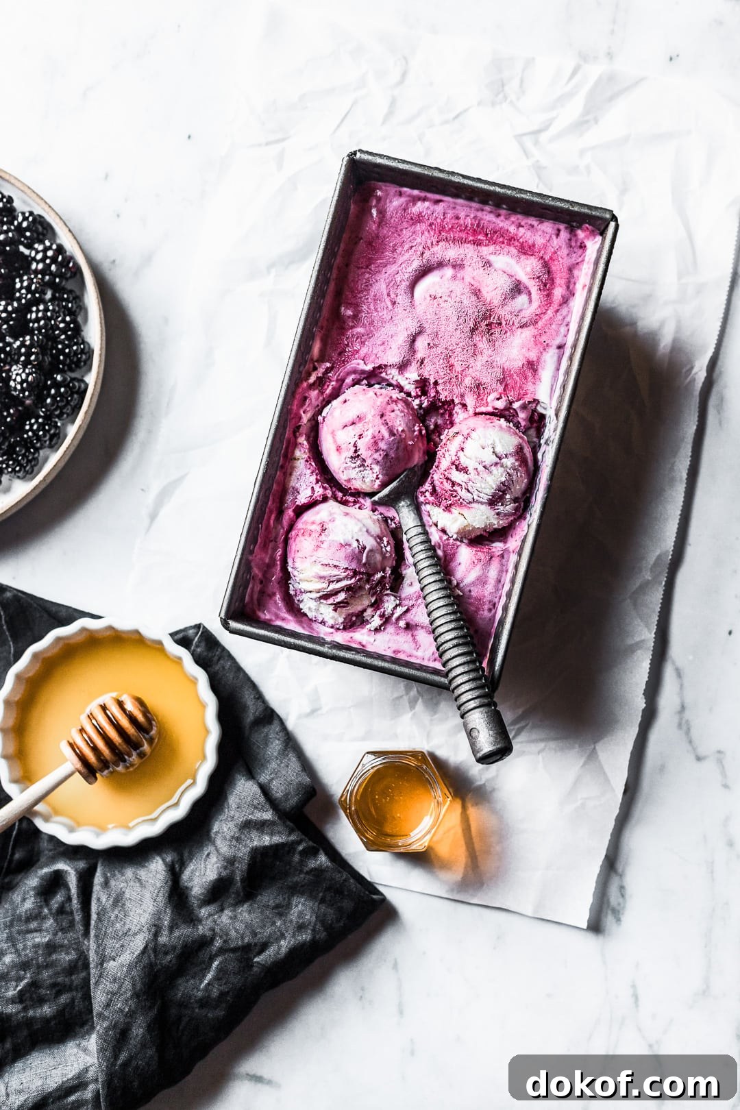 Three scoops of purple ice cream in container on a marble background with honey and blackberries nearby