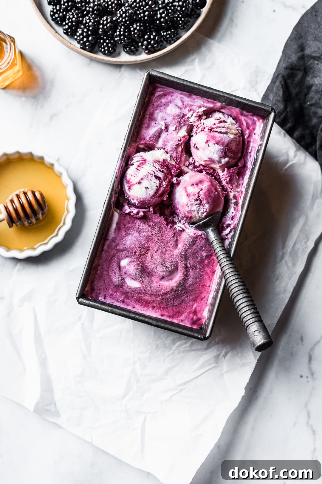 Three scoops of purple ice cream in container on a marble background with honey and blackberries nearby