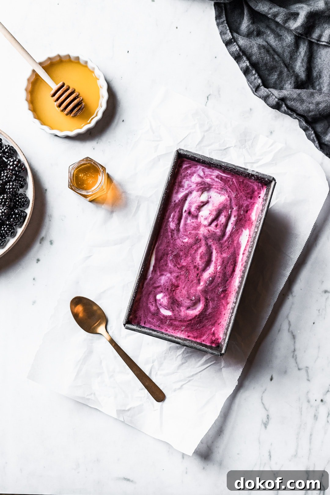 Purple and white swirled ice cream in a container on a marble background with blackberries and honey nearby