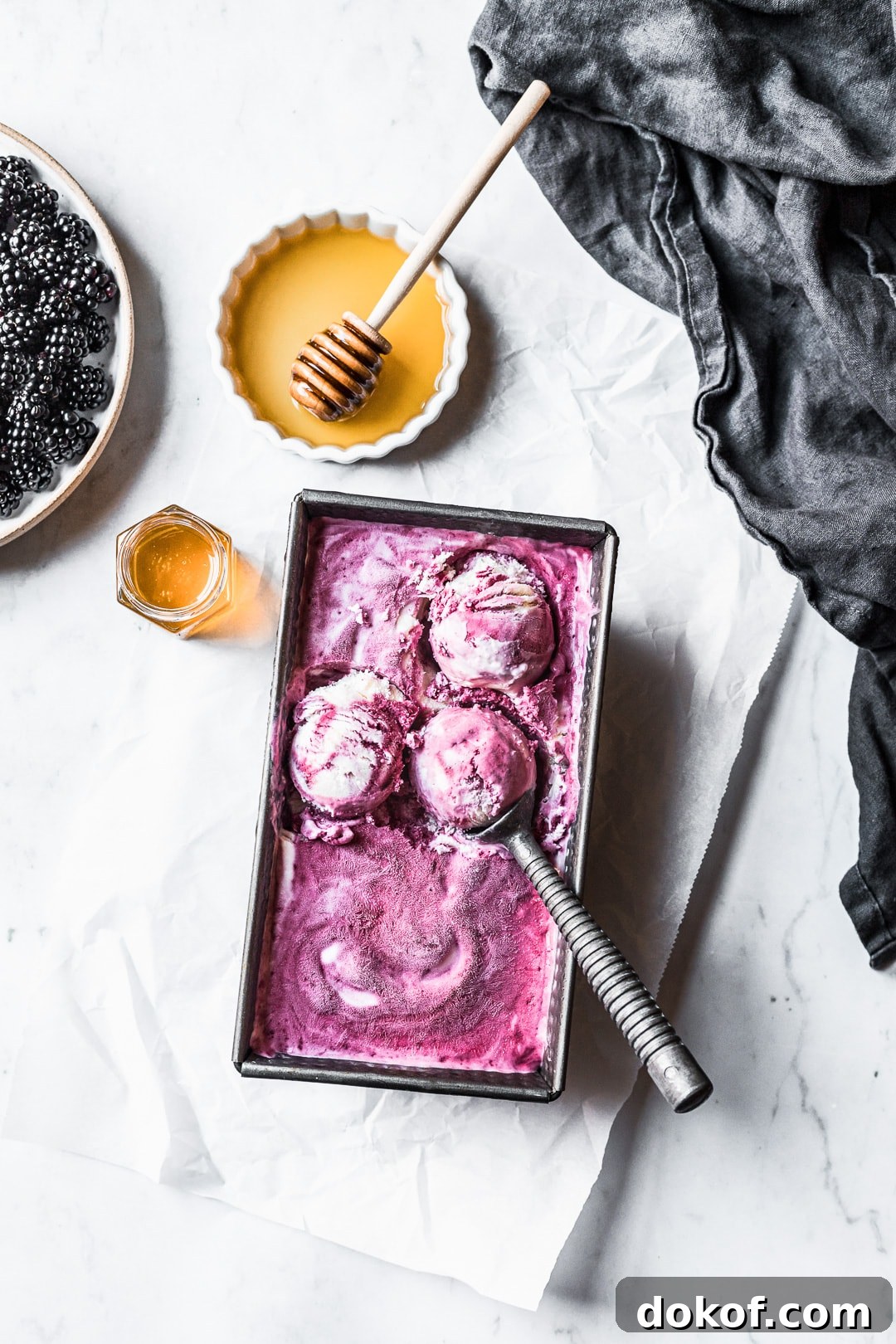 Three scoops of purple ice cream in container on a marble background with honey and blackberries nearby