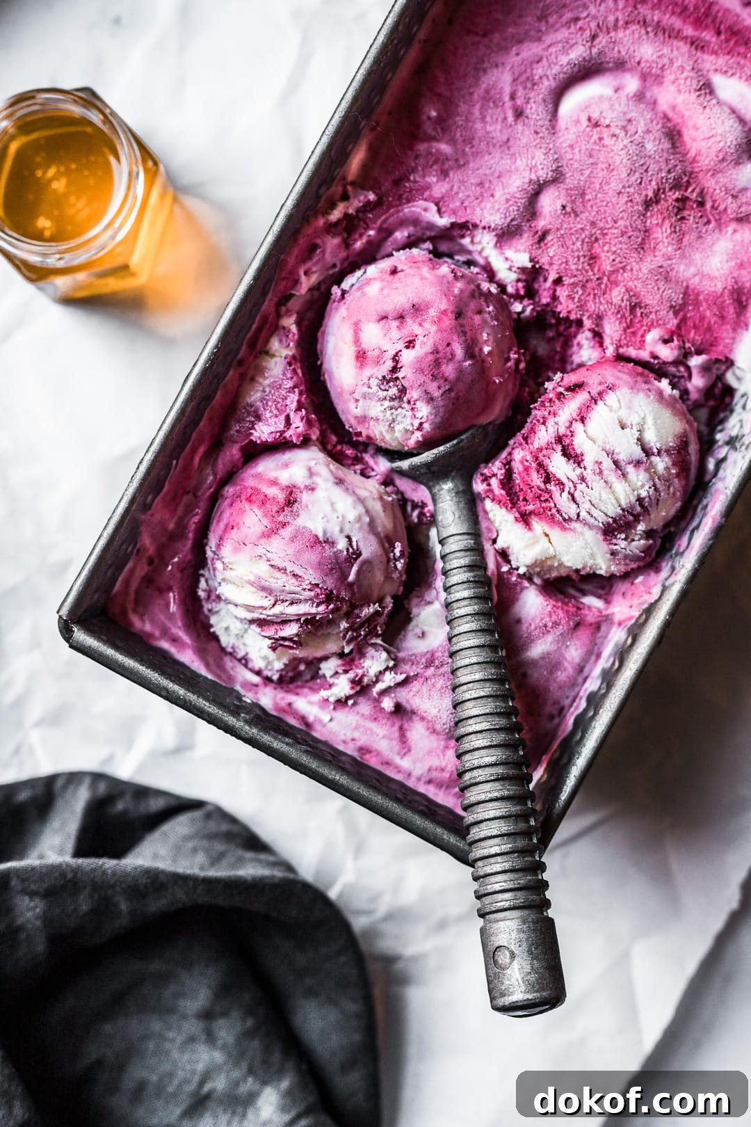Three scoops of purple and white ice cream in a container on parchment paper with honey nearby