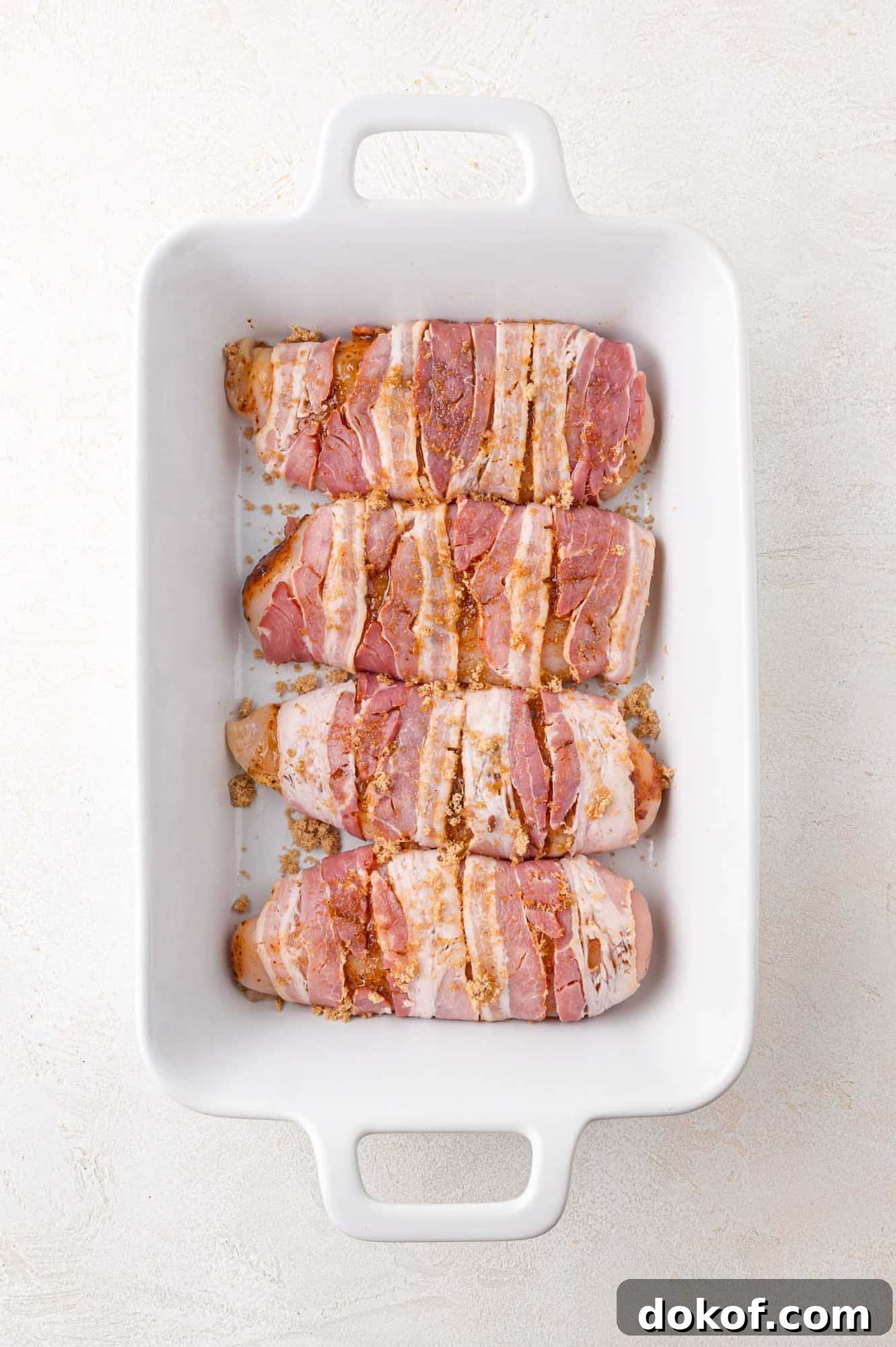 Juicy Bacon-Wrapped Chicken Breasts with Herb Butter Filling 6 Slices of seasoned bacon arranged in a white rectangular baking dish on a light surface.