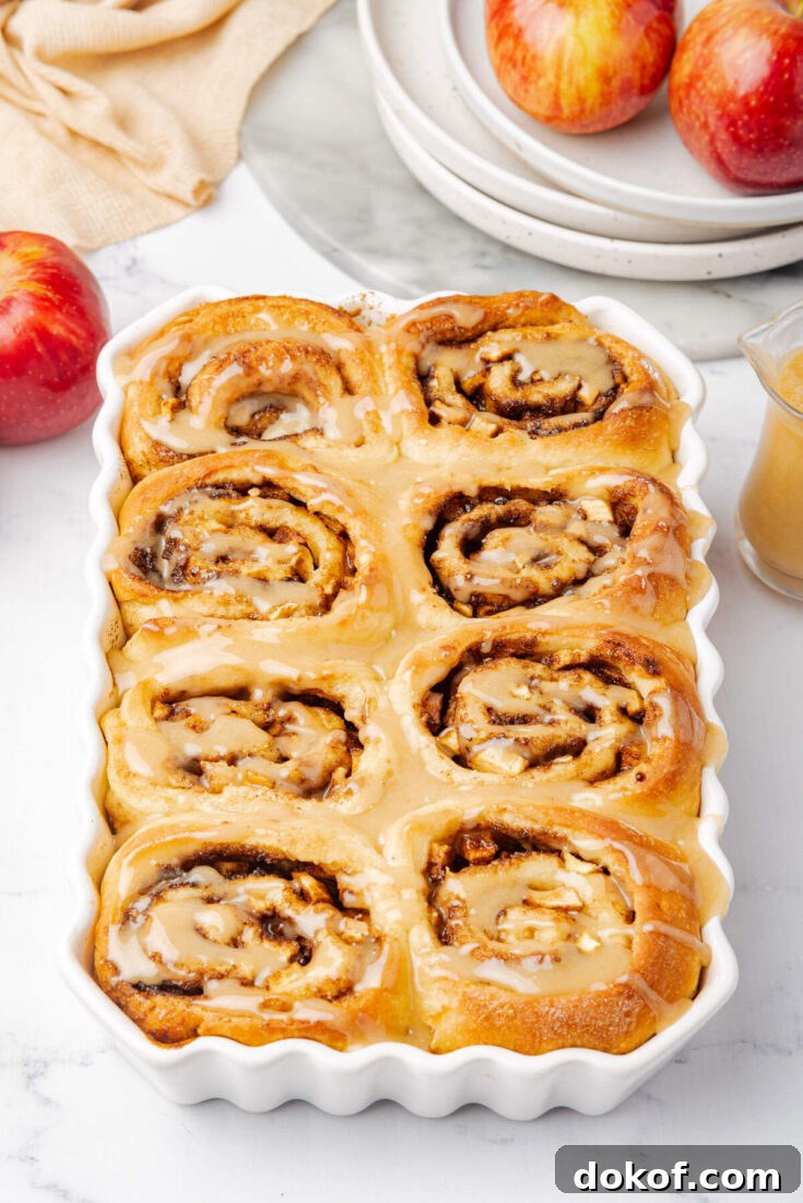 Overhead view of a full dish of warm cinnamon rolls, topped with a shiny glaze, with apples and plates nearby.