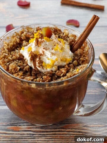 rhubarb crisp in a mug, topped with whipped cream with a stick of cinnamon, a spoon on the side and chopped rhubarb and cinnamon sticks strewn around