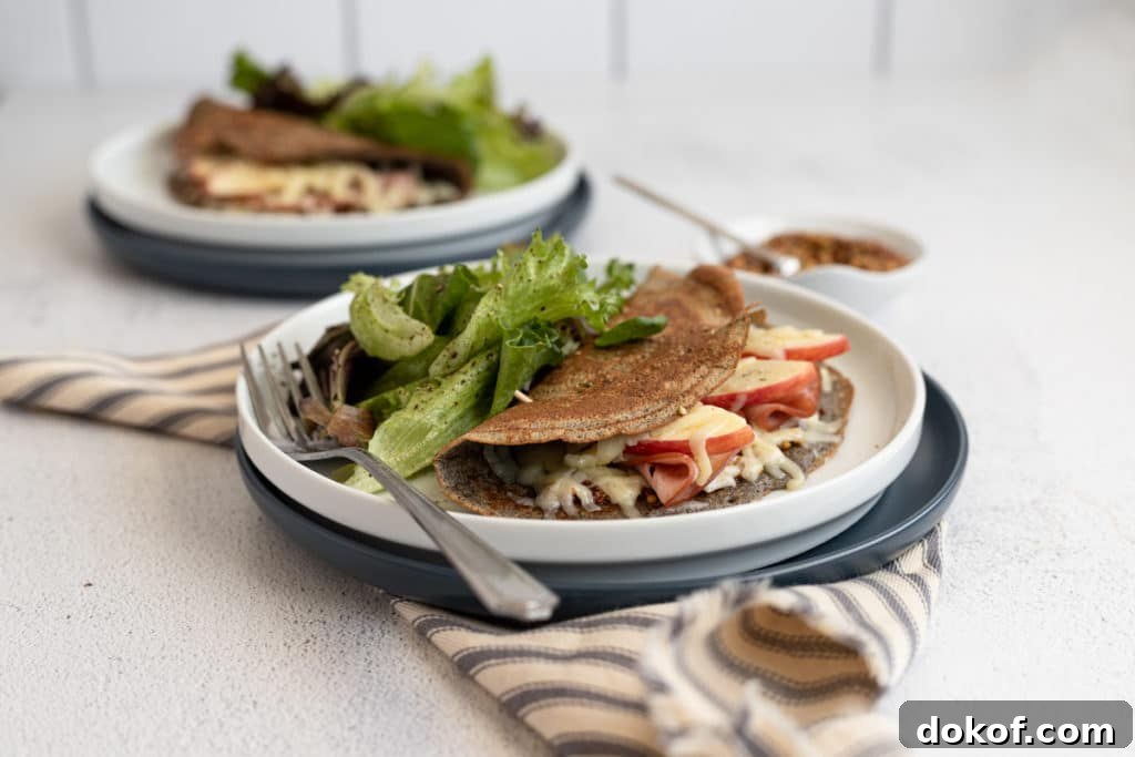 A buckwheat crepe on a plate filled with ham, cheese and apples, with a salad alongside.