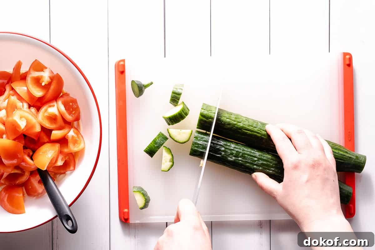 cucumber being cut into small chunks with bowl of tomatoes on the side