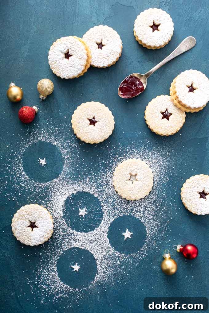 Classic Linzer Cookies Recipe: How to Make Austrian Jam Sandwich Cookies 5 linzer cookies over a blue surface with confectioners' sugar all over.
