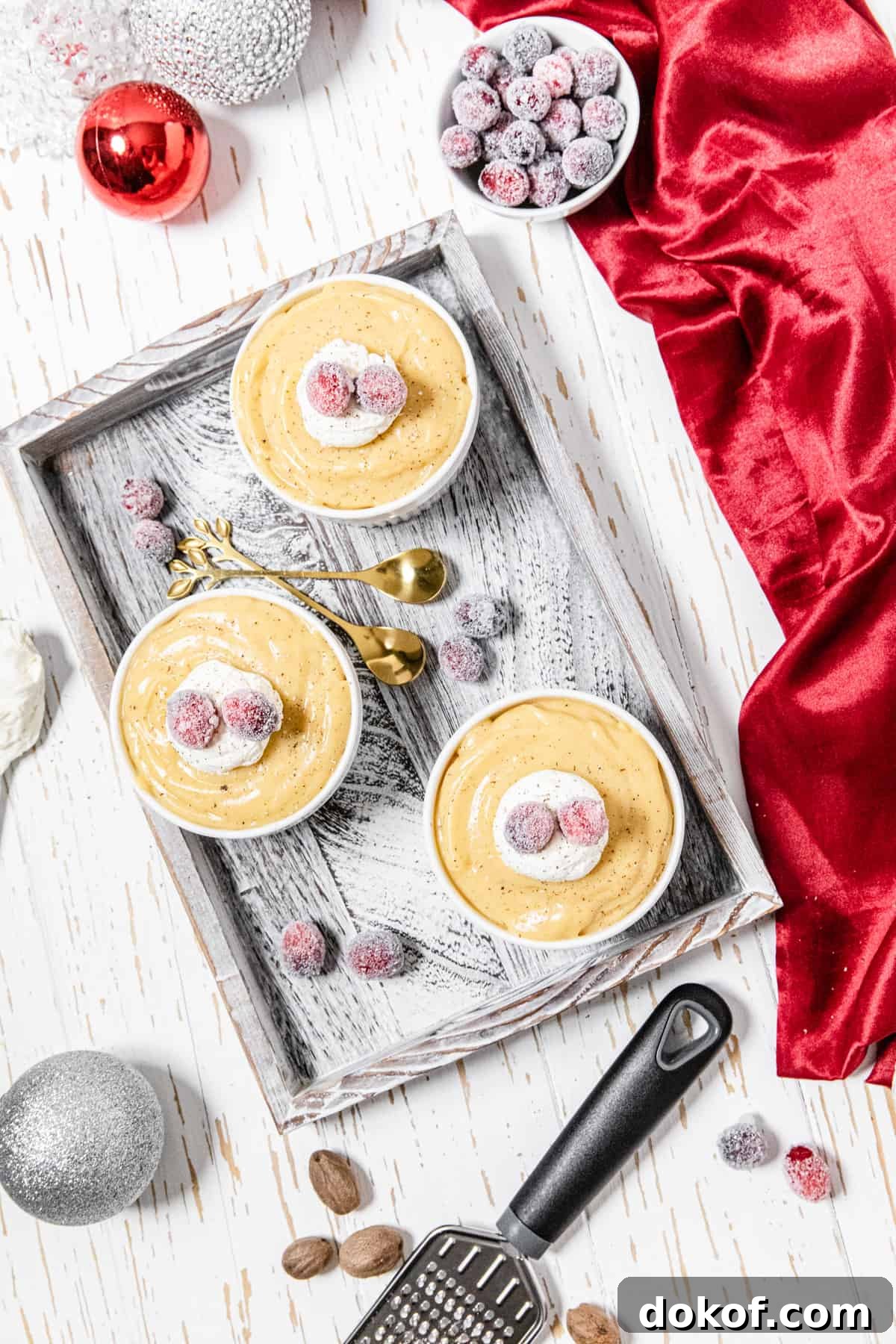 Creamy Homemade Eggnog Pudding Recipe for the Holidays 6 flat-lay of three ramekins with pudding on a wood tray with sugared cranberries, whole nutmegs and red and white ornaments around it