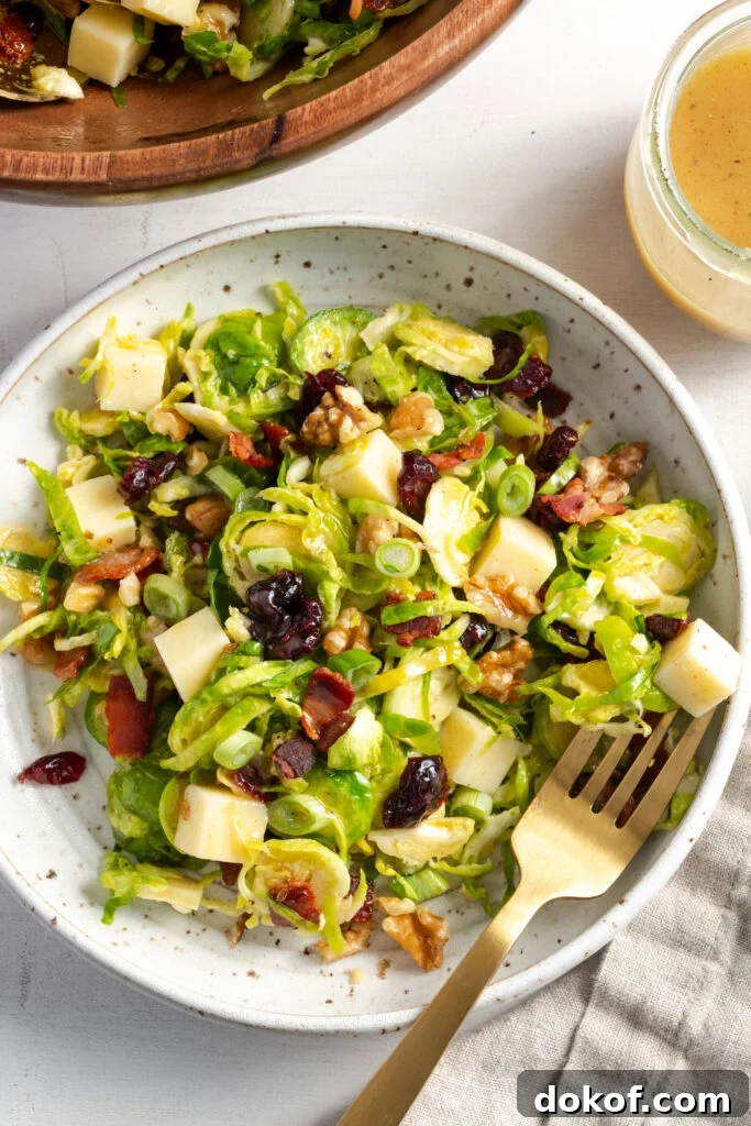 Citrus-Shaved Brussels Sprout Salad with Toasted Almonds 2 overhead shot of a flecked white earthenware pasta bowl filled with a portion of shaved brussel sprouts salad with bacon, cheese, and cranberries on a white table with a gold fork.