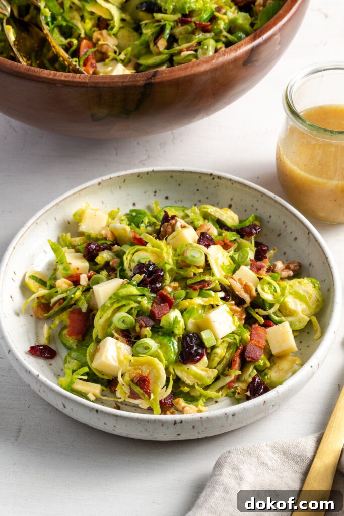 Citrus-Shaved Brussels Sprout Salad with Toasted Almonds 10 45 degree angle shot of a white flecked earthenware shallow bowl filled with a serving of shaved brussel sprouts salad on a white table with the wooden serving bowl and a small glass jar with extra vinaigrette.