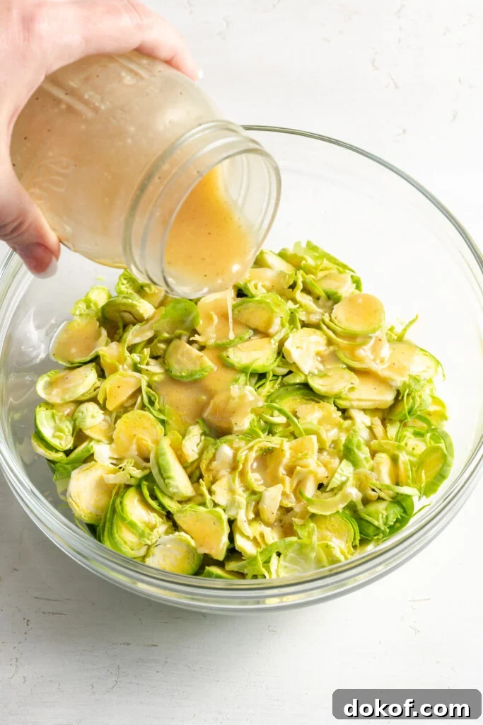 Citrus-Shaved Brussels Sprout Salad with Toasted Almonds 8 hand pouring vinaigrette from a mason jar over the shaved brussels sprouts in the bowl.