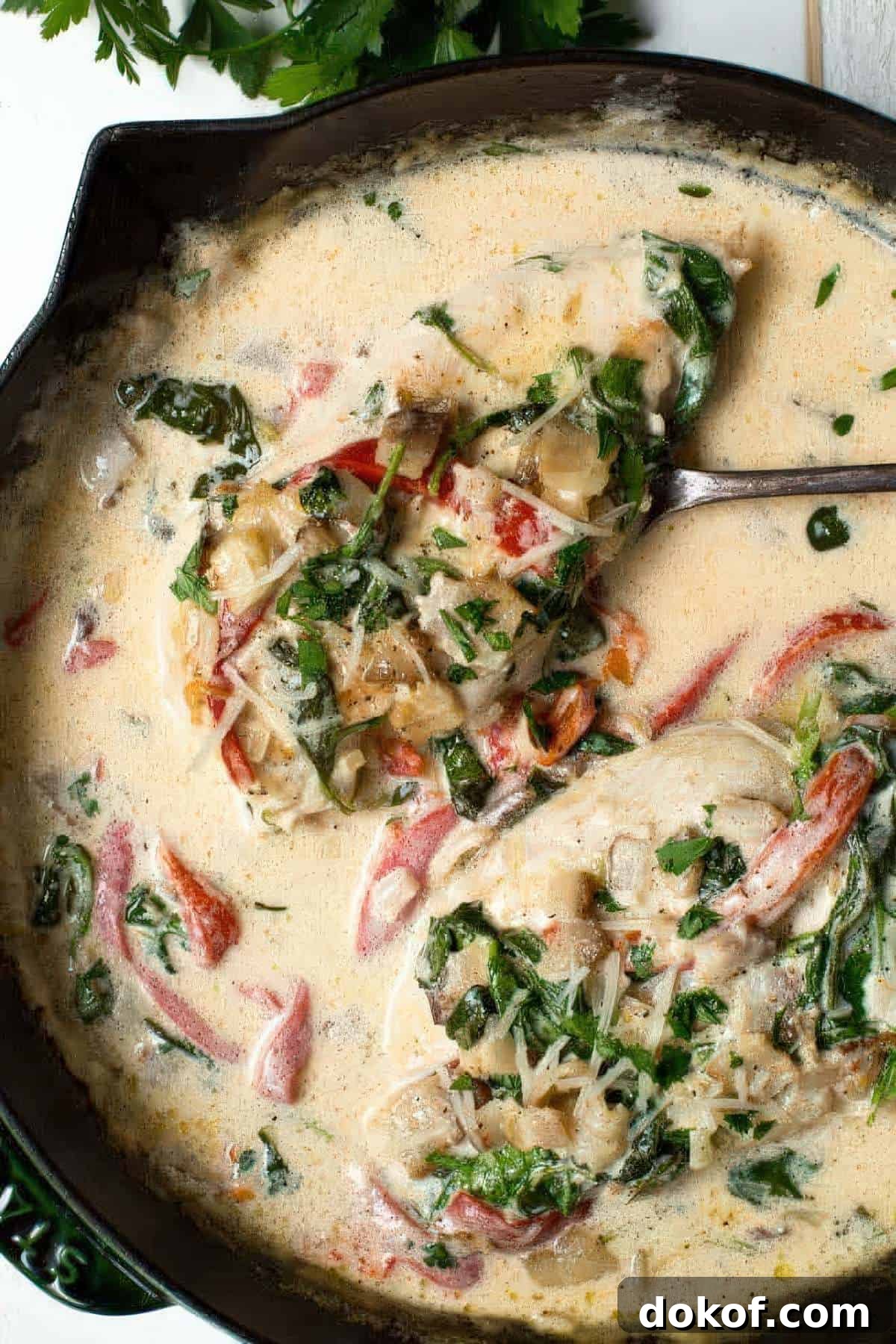 Spoon lifting creamy chicken breast from skillet with spinach, roasted red peppers, and parmesan in a garlic cream sauce