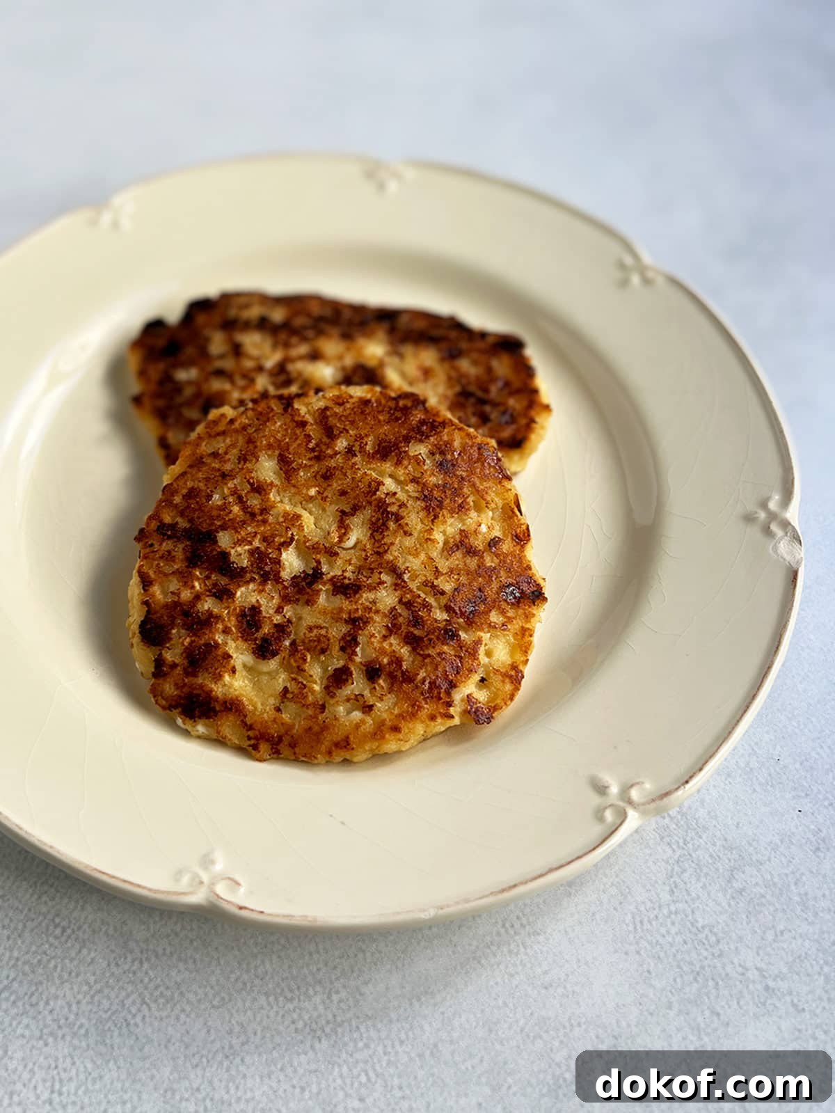 Two cheese latkes on an off white plate with a close up of the top of one of the pancakes.