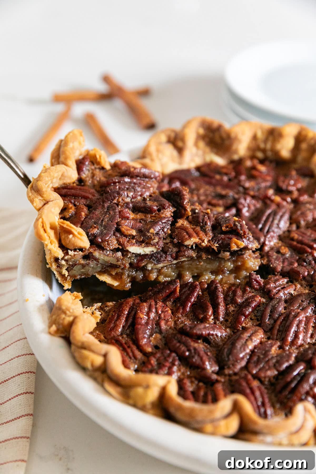 A slice of pecan pie being pulled from the pie with plates and cinnamon sticks in the background.