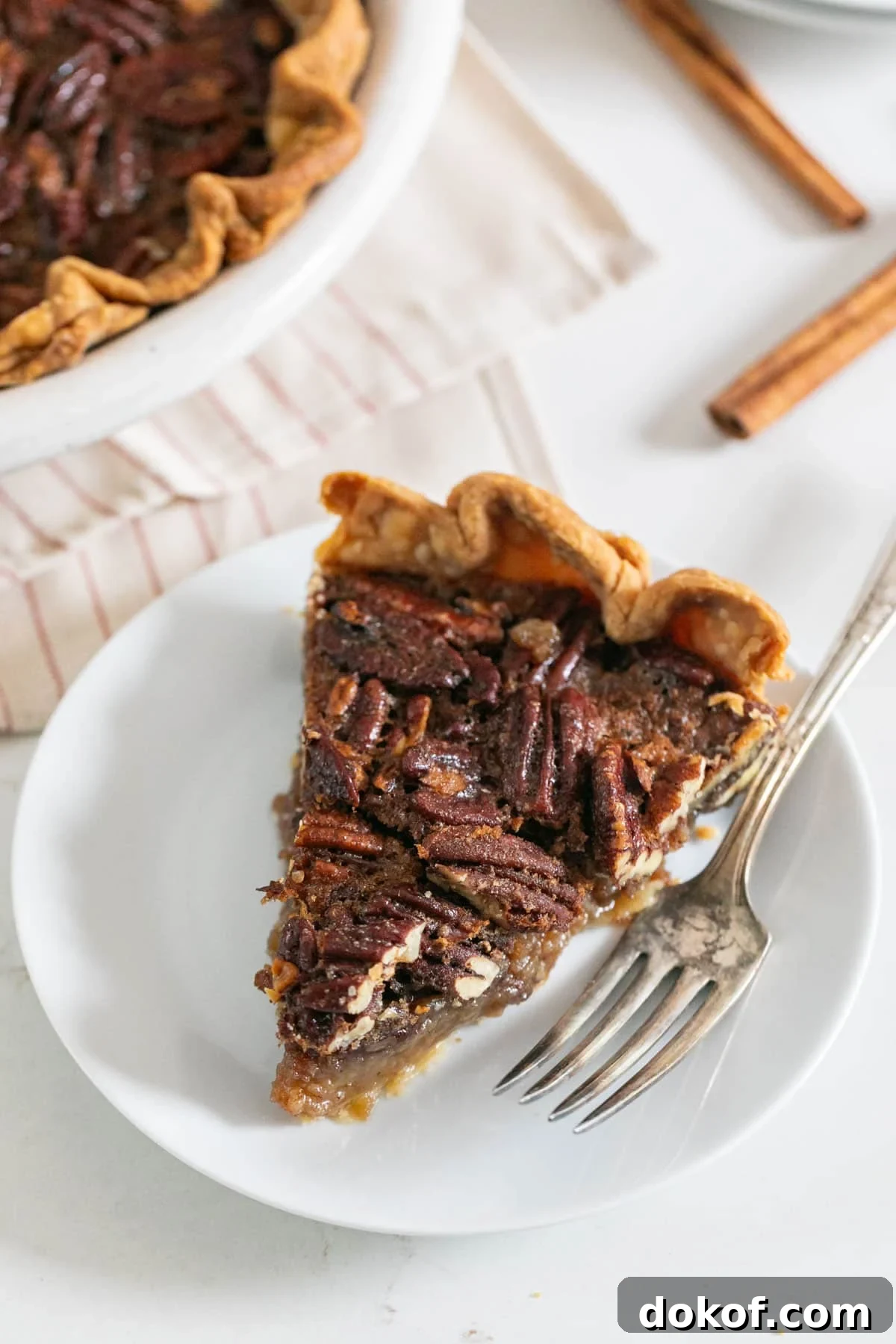 A slice of pecan pie on a white plate with a fork on it and the rest of the pie behind it.