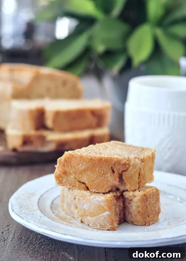 one slice of snickerdoodle bread sliced in half and stacked, the remaining full loaf in blurred background next to a white ceramic mug.