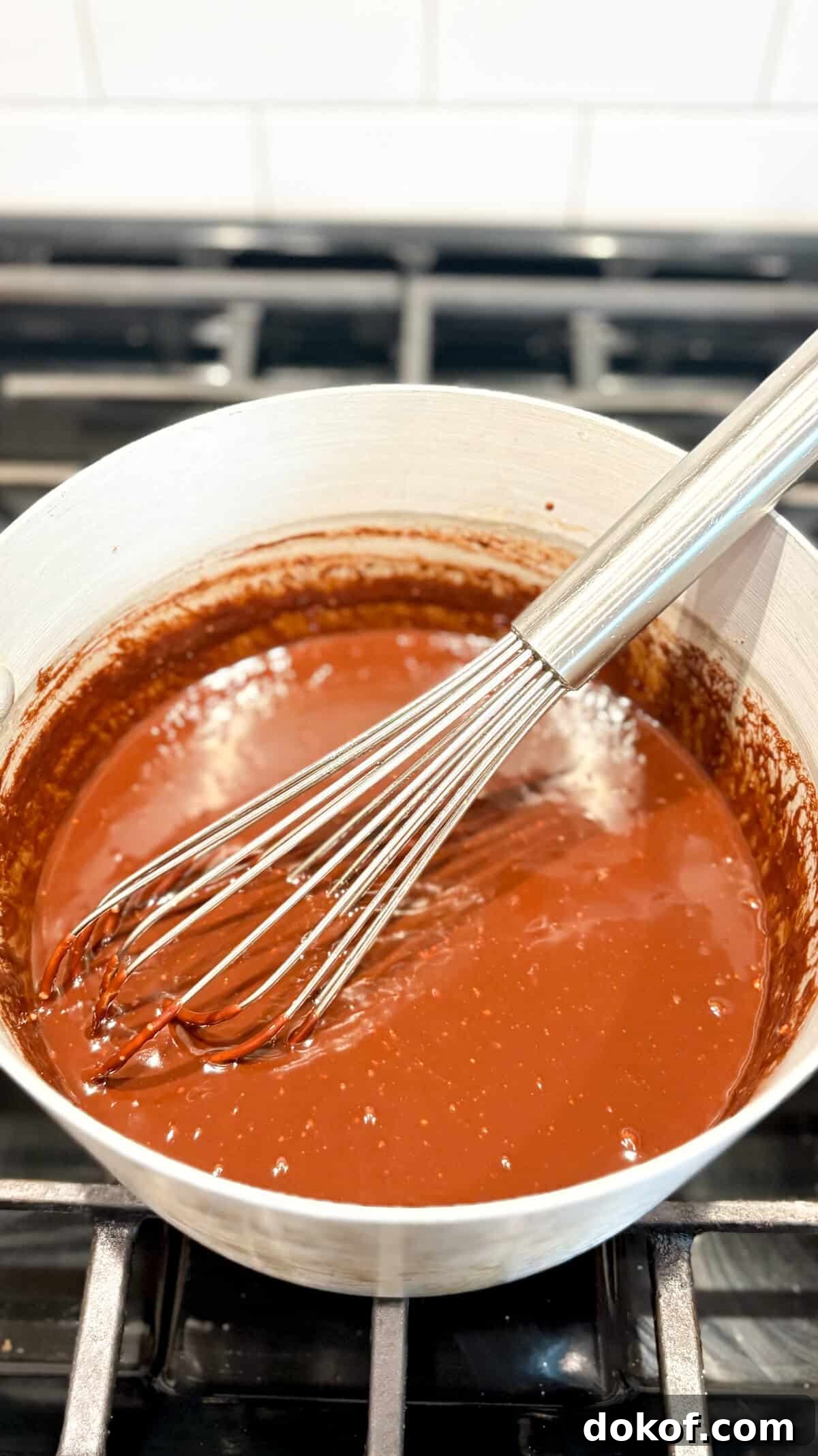 Chocolate ganache frosting in a pot on the stove, with a whisk.