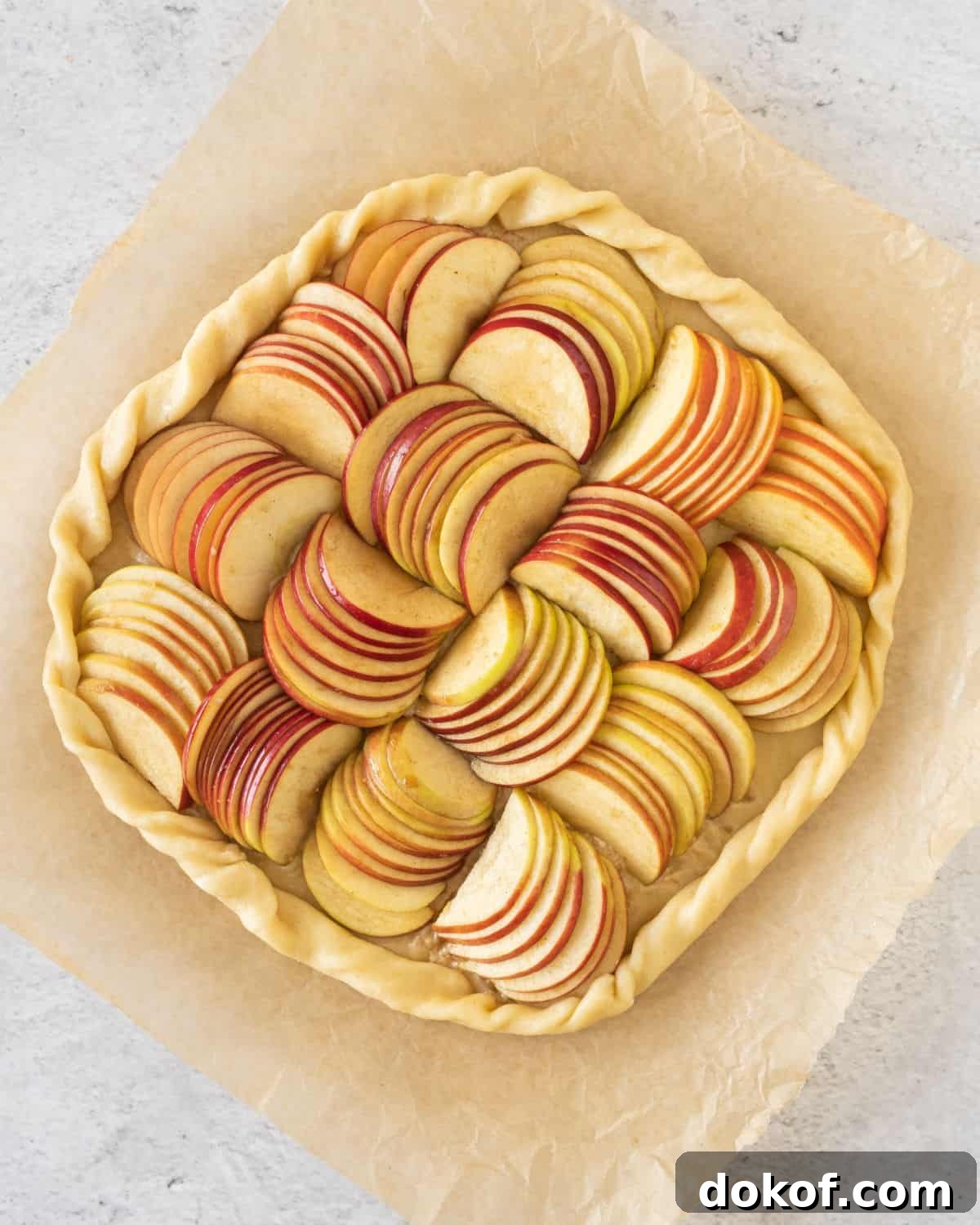 Rustic Apple Galette Recipe: Simple Homemade Apple Tart 7 Sliced apples arranged over pie crust to make an apple galette. Beige parchment paper, grey surface.