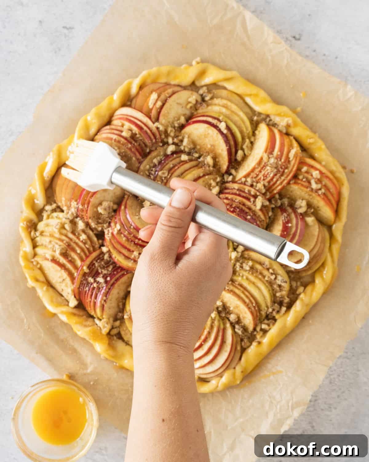 Rustic Apple Galette Recipe: Simple Homemade Apple Tart 8 Brushing egg wash on an assembled rustic apple tart on a beige parchment paper.