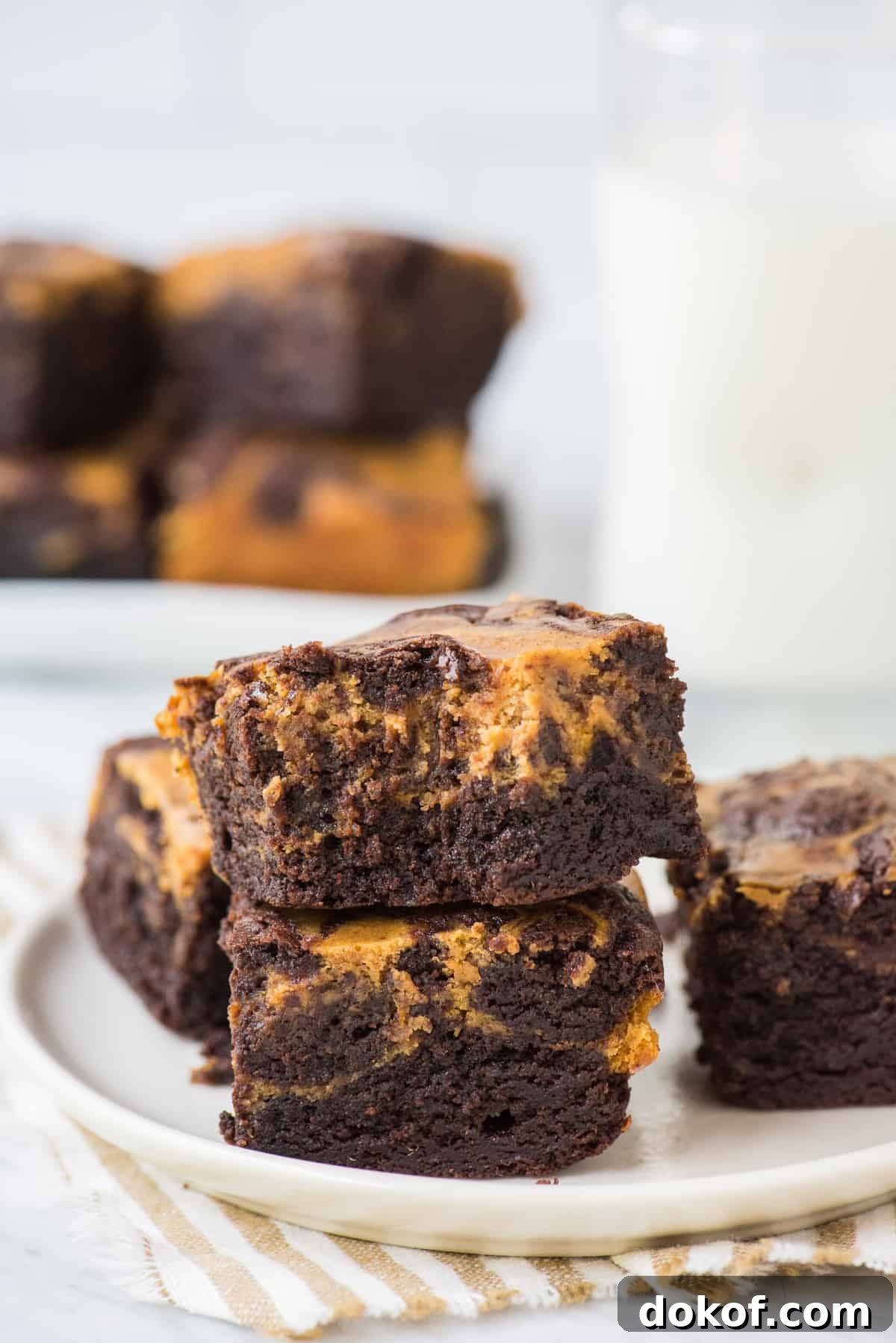 2 pumpkin cheesecake brownies stacked on top of each other on a round cream plate with striped linen underneath the plate