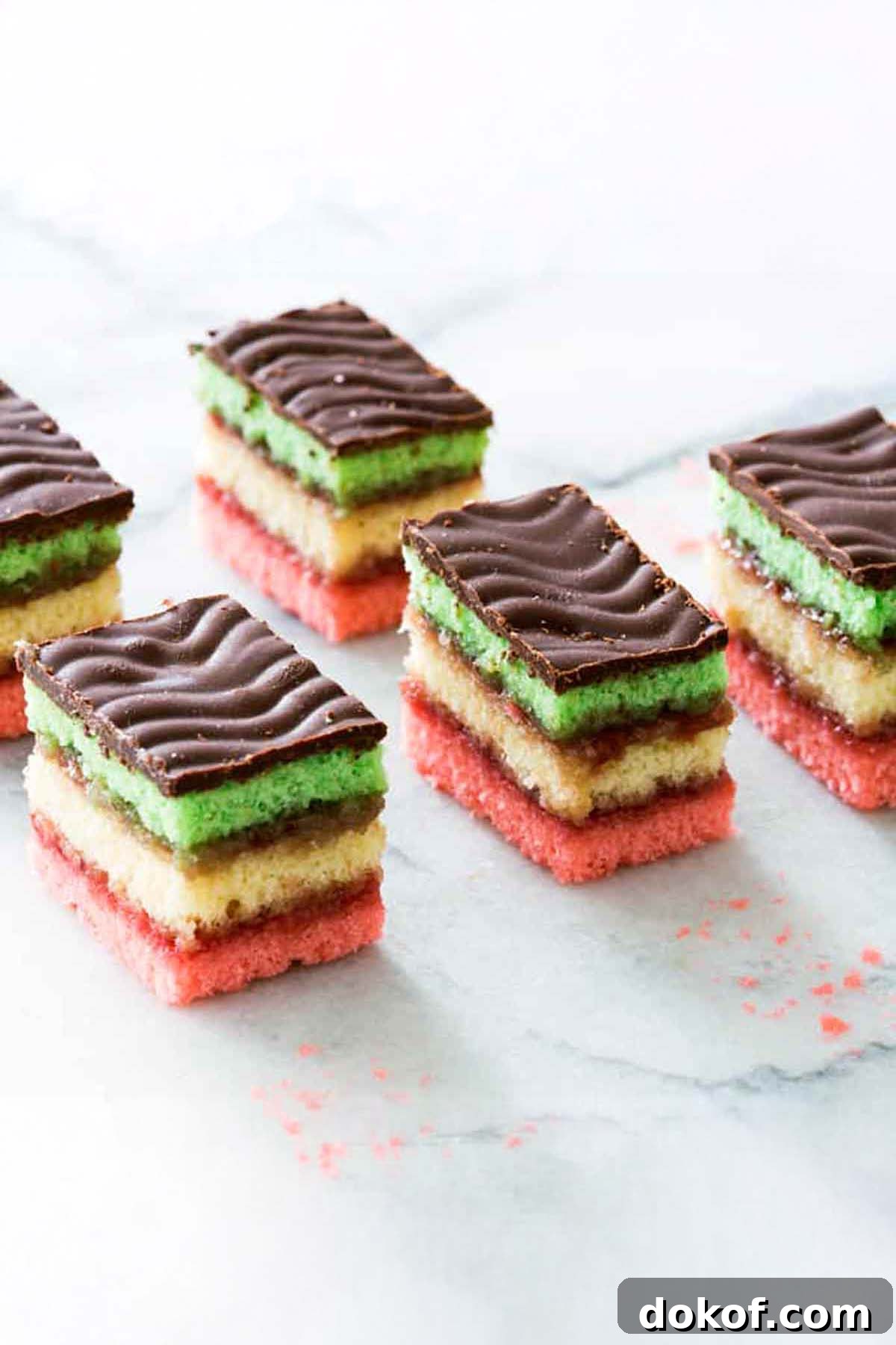 Five squares of Italian rainbow cookies, with layers of pink, yellow, green cake layers and a chocolate topping.