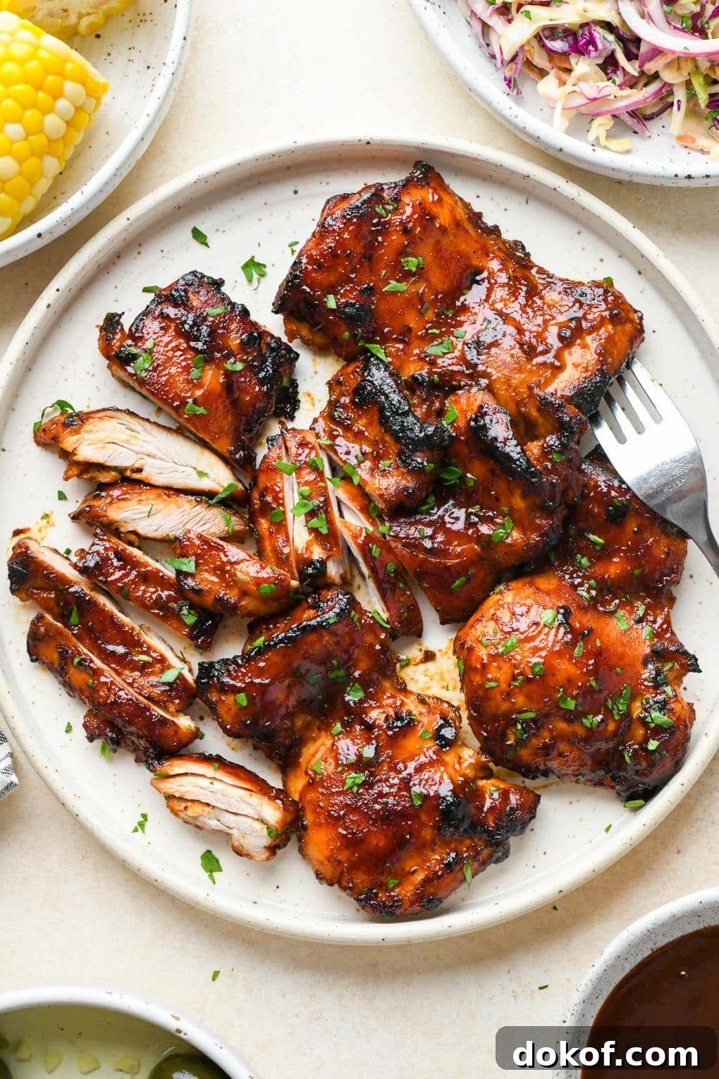 Oven-Baked Caramelized BBQ Chicken Thighs with Sticky Glaze 14 A large white ceramic plate of boneless skinless bbq chicken thighs topped with chopped parsley, next to a bowl of coleslaw and some corn on the cob.