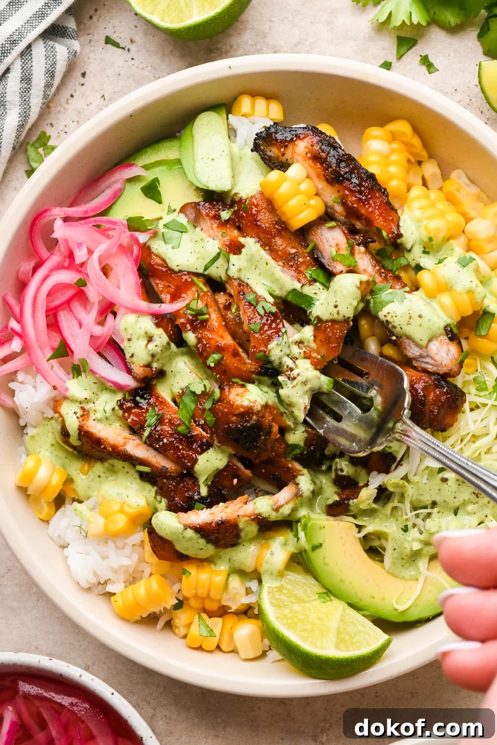 Oven-Baked Caramelized BBQ Chicken Thighs with Sticky Glaze 15 BBQ chicken rice bowl filled with rice, cooked sweet corn, thinly shredded cabbage, avocado, pickled red onions, and sliced BBQ chicken thighs. Drizzled with a bright green cilantro ranch dressing and garnished with chopped cilantro. A fork is digging into the chicken.