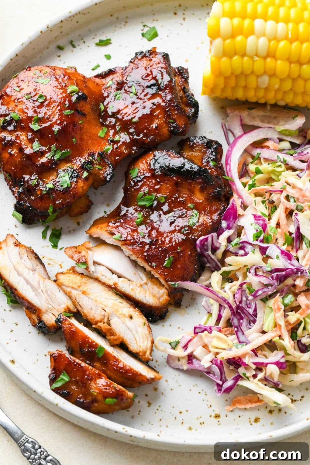 Oven-Baked Caramelized BBQ Chicken Thighs with Sticky Glaze 16 A large white ceramic plate of boneless skinless bbq chicken thighs next to corn on the cob and creamy coleslaw.