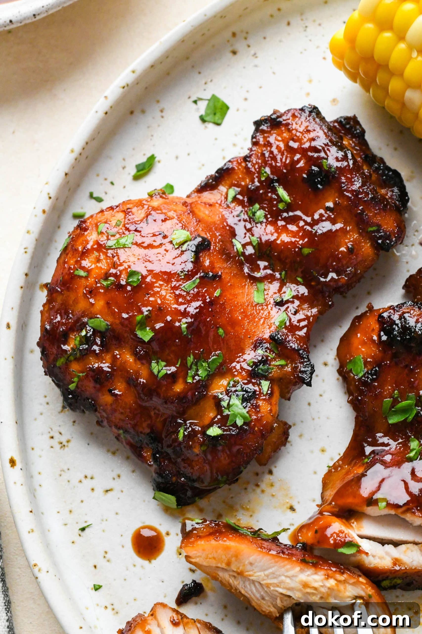 Oven-Baked Caramelized BBQ Chicken Thighs with Sticky Glaze 17 A single piece of BBQ chicken thigh perfectly glazed and glistening on a white speckled plate, next to corn on the cob and some creamy coleslaw.