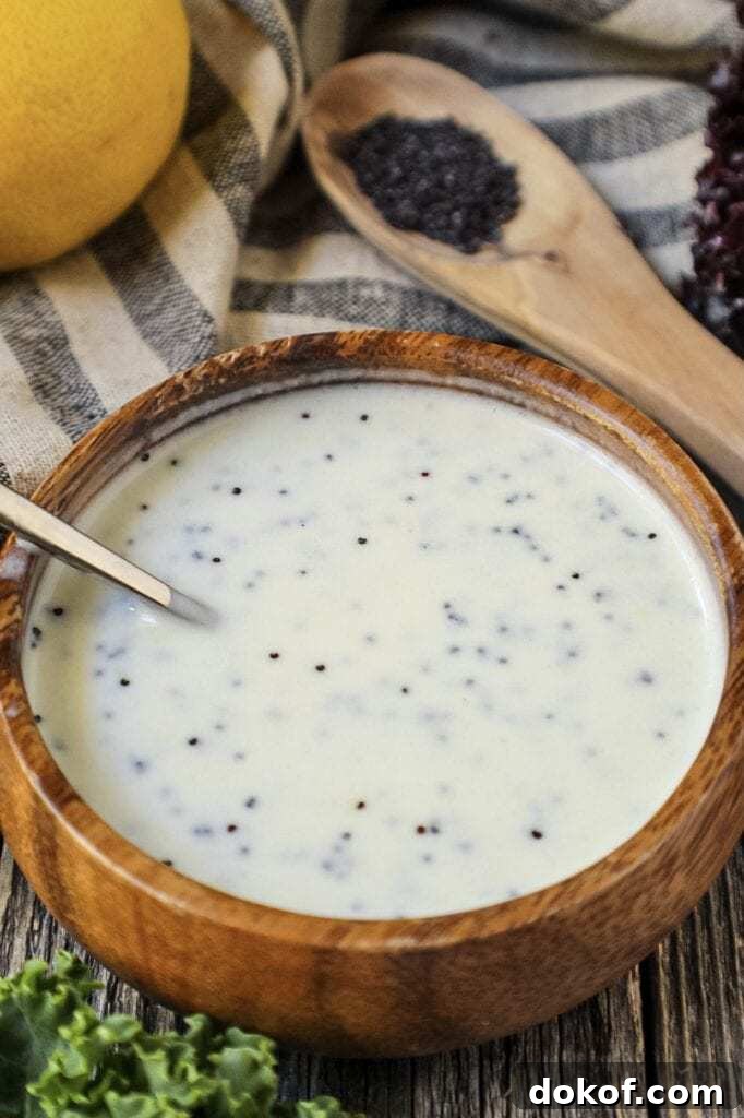 Creamy Lemon Poppyseed Dressing in a wooden bowl with wooden spoon filled with poppy seeds in shot along with a whole lemon, leaf lettuce and cloth napkin
