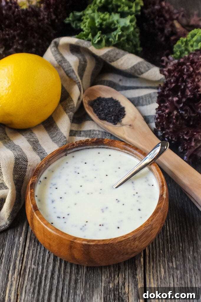 Creamy Lemon Poppyseed Dressing in a wooden bowl with wooden spoon filled with poppy seeds in shot along with a whole lemon, leaf lettuce and cloth napkin