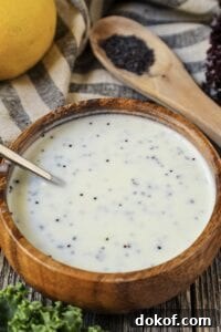 Creamy Lemon Poppyseed Dressing in a wooden bowl with wooden spoon filled with poppy seeds in shot along with a whole lemon, leaf lettuce and cloth napkin