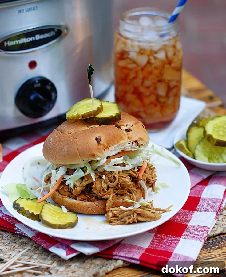 A Shredded Chicken BBQ Sandwich with pickles and a crock pot in the background.