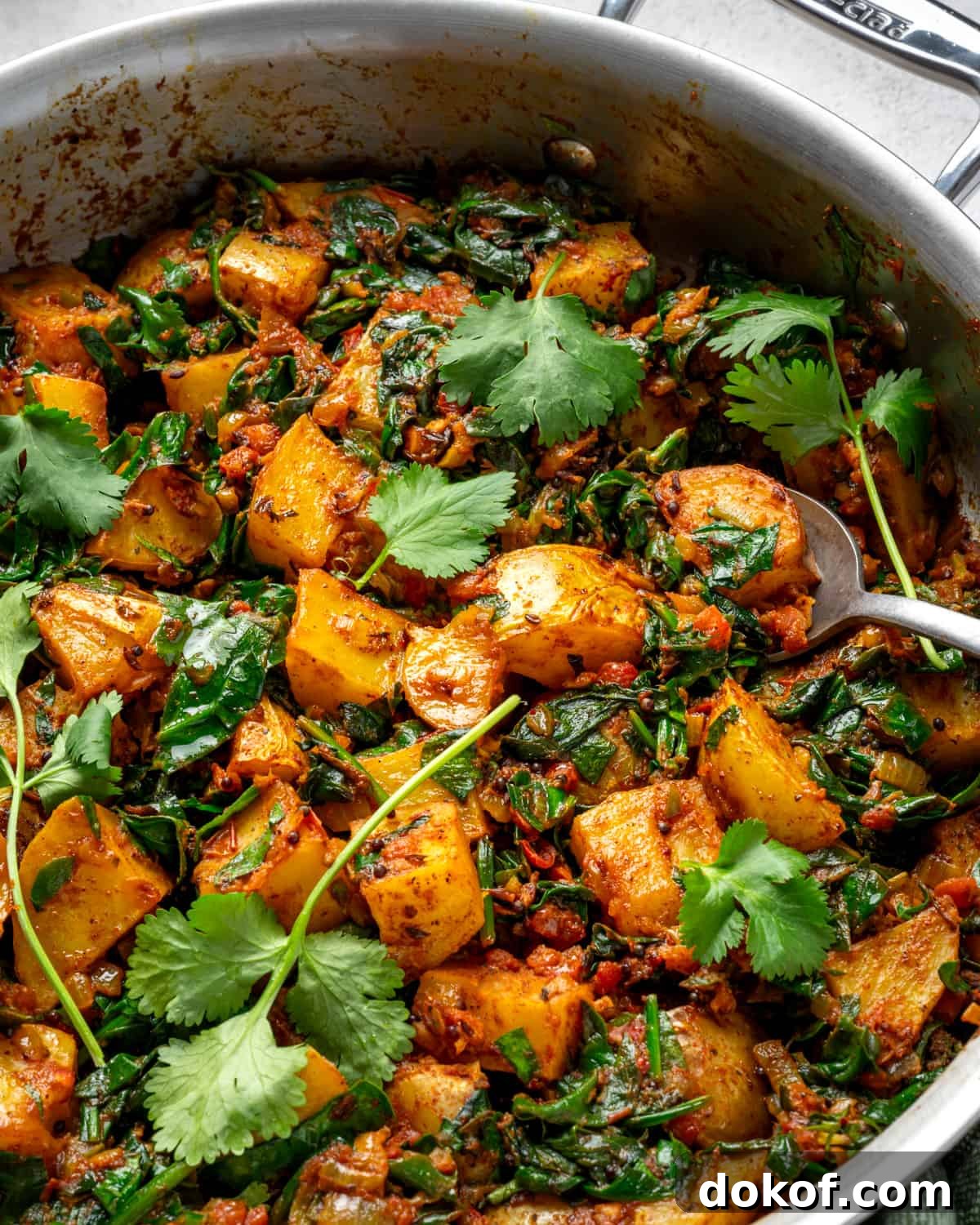 close up photo of saag aloo in saucepan with cilantro leaves and a spoon.