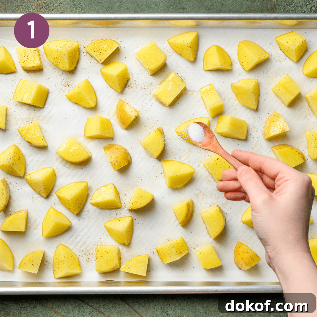 person sprinkling salt over cubed potatoes on a lined baking tray.