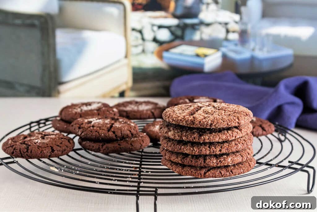 A round, black wire cooling rack with cocoa cookies cooling on it on a cream-colored surface with living room furniture in the background.