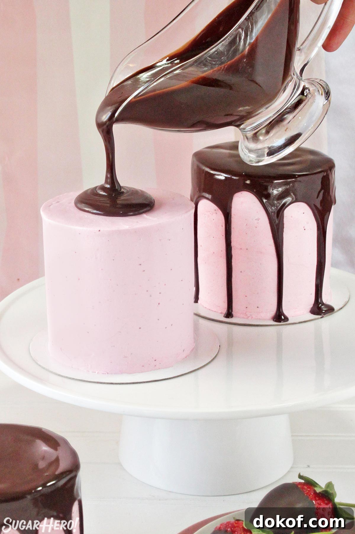 Pouring shiny chocolate ganache on top of a pink-frosted miniature cake.