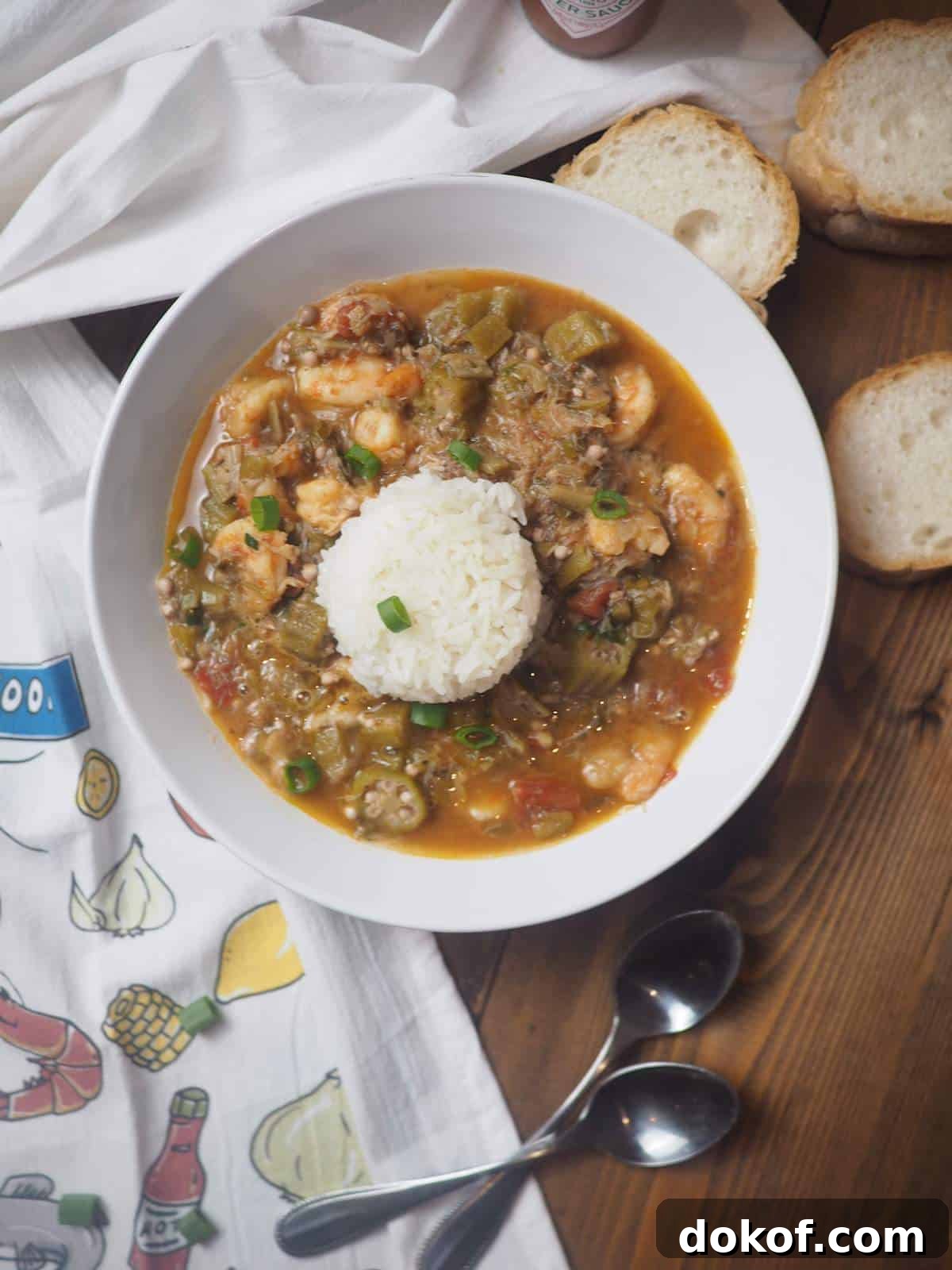 A white bowl of seafood gumbo and rice with two spoons and french bread.