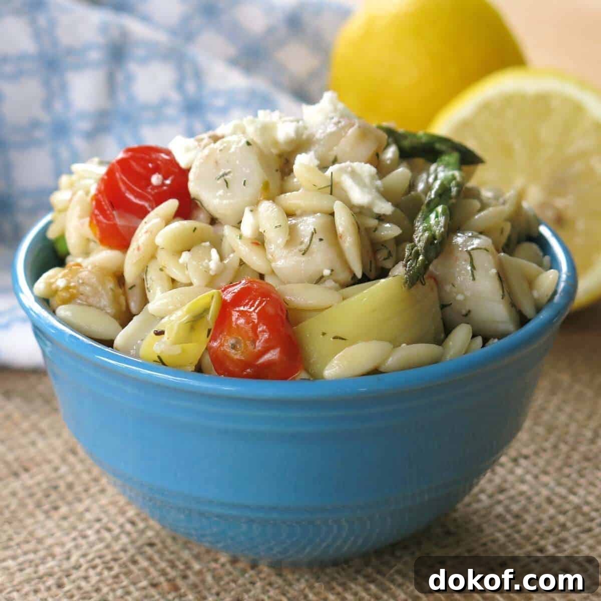 Warm orzo salad with scallops, artichokes, tomatoes, asparagus, and feta in a bowl with lemons behind it.