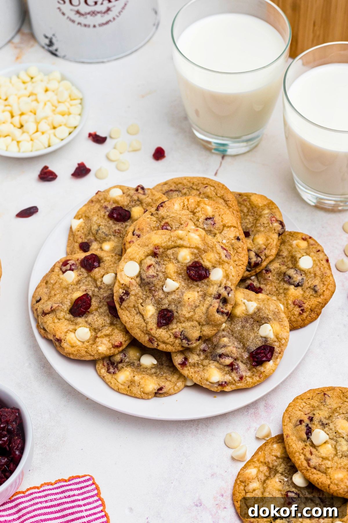 White chocolate chip and cranberry cookies stacked on a white plate with glasses of milk on the table. 