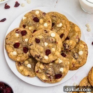 White chocolate chip and cranberry cookies stacked on a white plate with glasses of milk on the table.